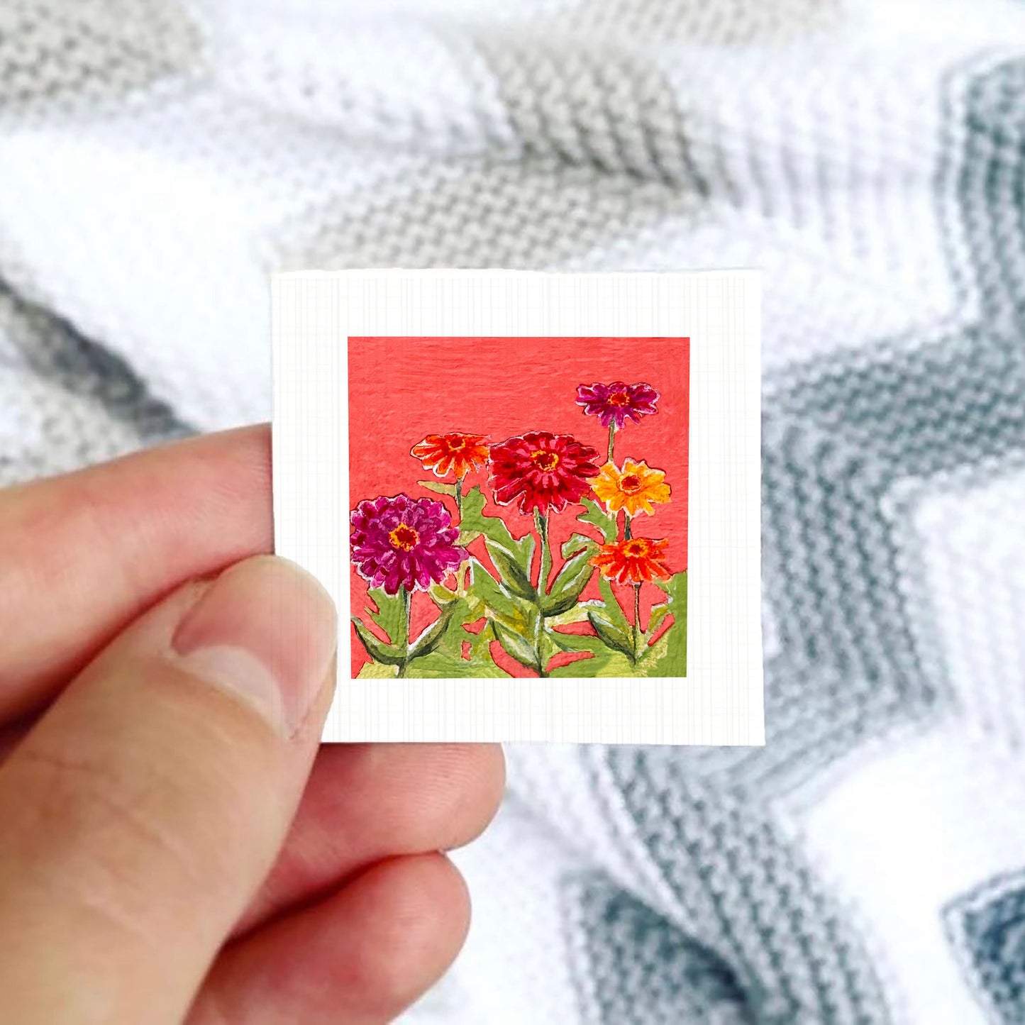 A hand is holding a small square card with a vibrant painting of colorful flowers against a red background.
