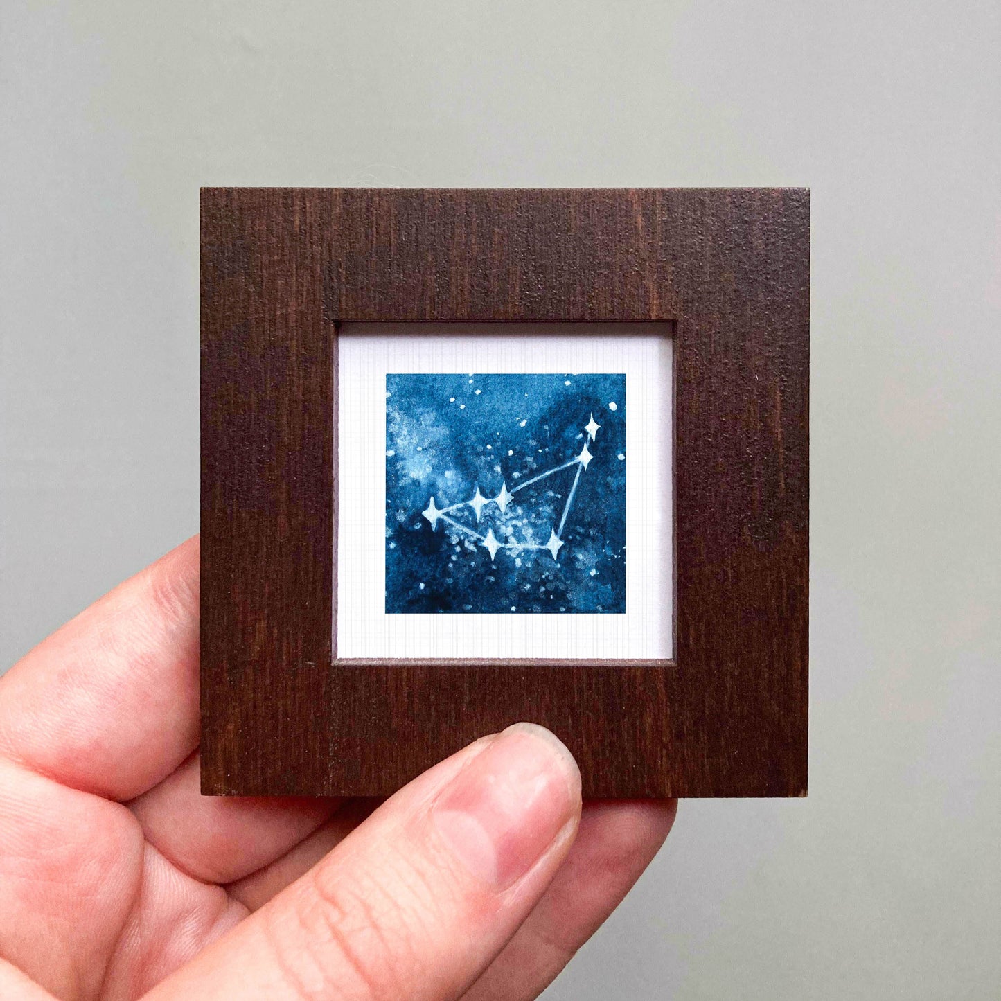 A hand is holding a small wooden frame containing a framed image of a starry night sky.