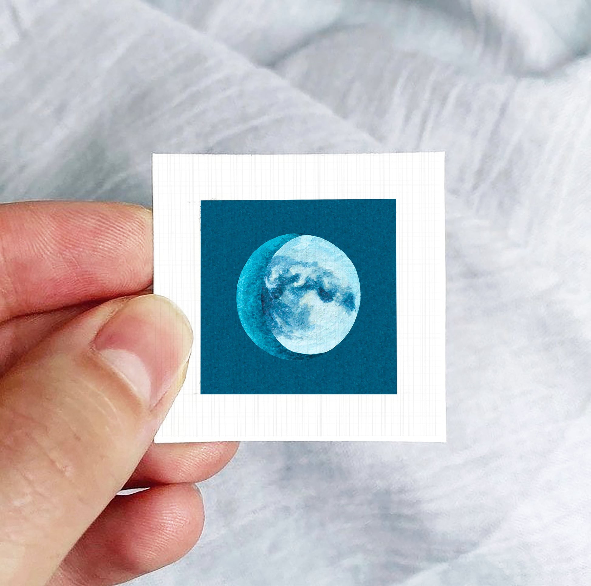 A hand is holding a small square-shaped paper with a depiction of a crescent moon on it.