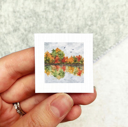 A hand holding a small square picture depicting a landscape with trees and a body of water.
