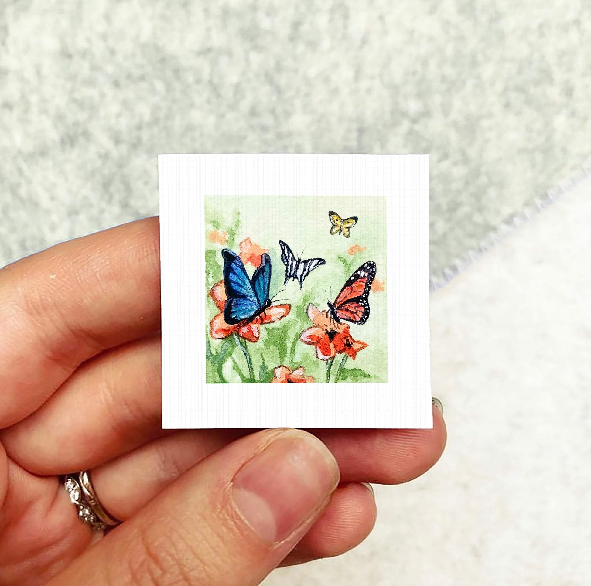 A hand holding a small square artwork depicting colorful butterflies on flowers.