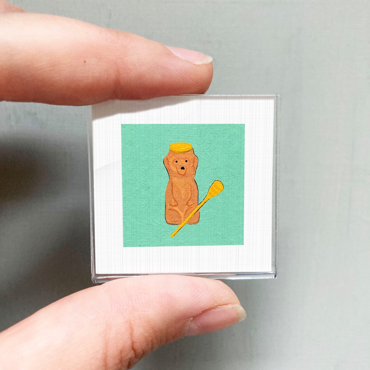 A person's hand is holding a small square with a picture of a brown bear wearing a yellow hat and holding a paddle.