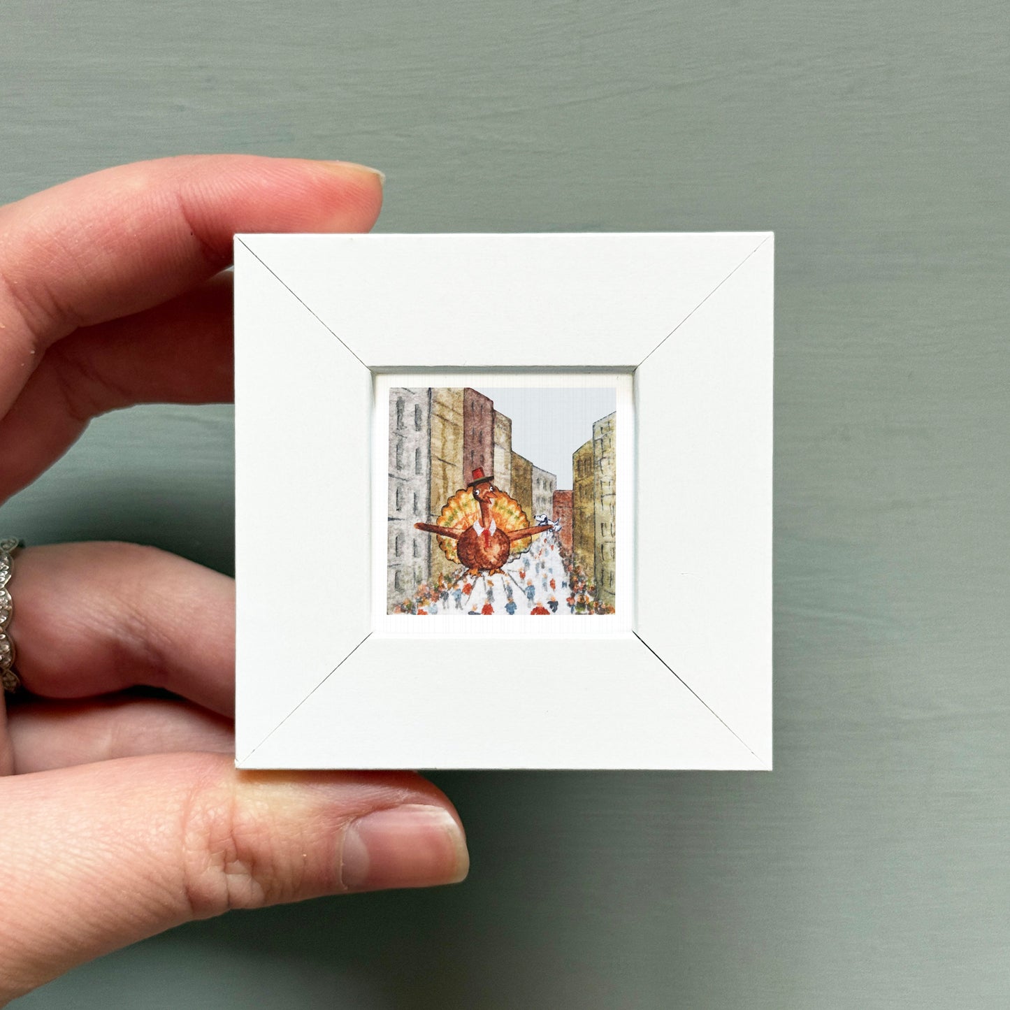 A hand is holding a small white square frame containing a miniature painting of a person in a colorful outfit.