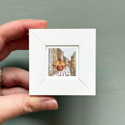 A hand is holding a small white square frame containing a miniature painting of a person in a colorful outfit.