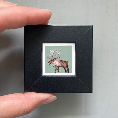 A person's hand is holding a small black picture frame containing a framed image of a deer with large antlers.