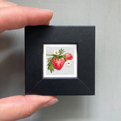 A person's hand holding a small black frame containing a framed image of a strawberry with white flowers.