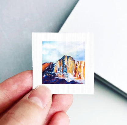 A hand is holding a small square-shaped piece of paper with a painting of a mountain landscape on it.