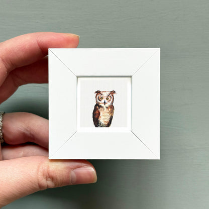 A hand is holding a small white square frame containing a painting of an owl.