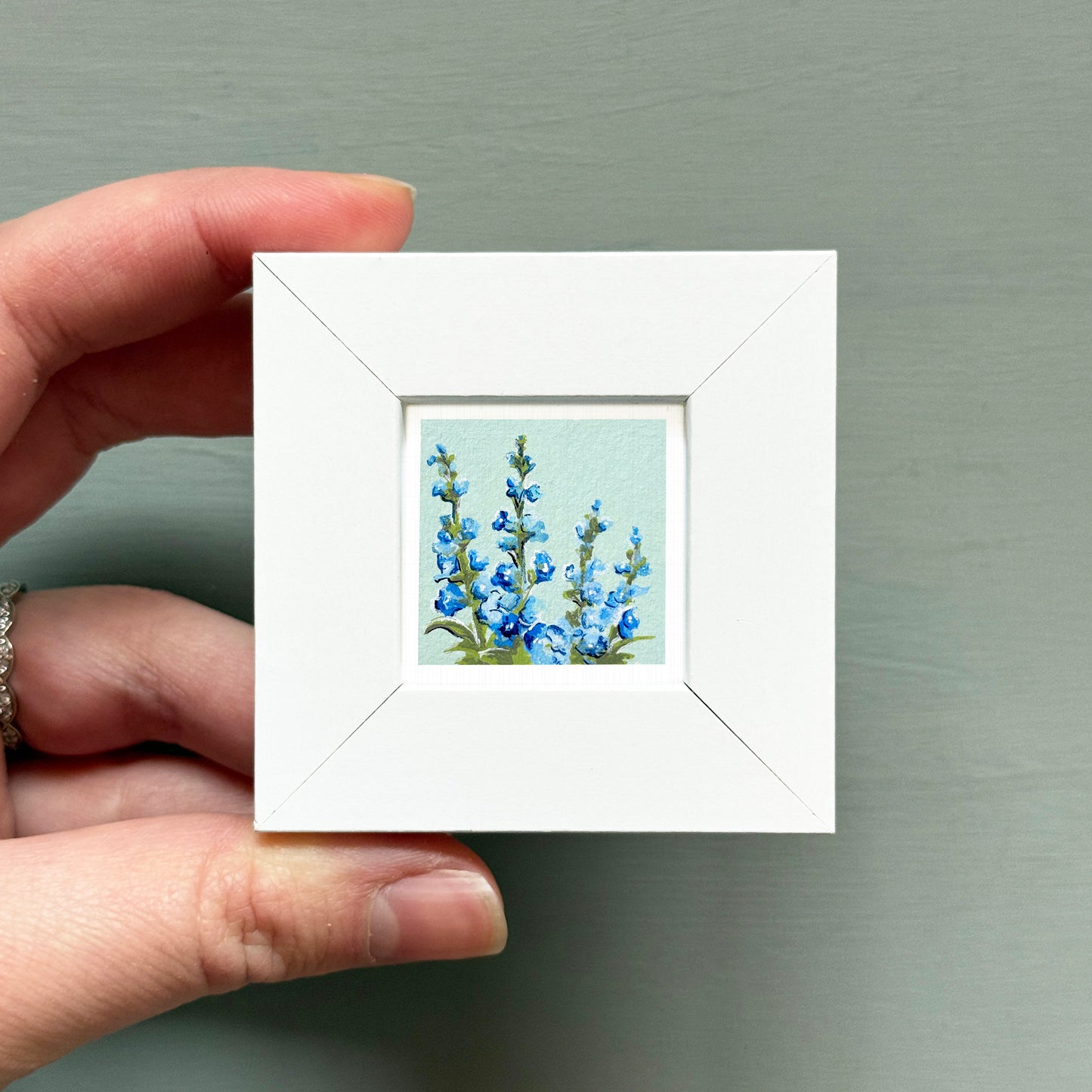 A hand is holding a small white square frame containing a painting of blue flowers.