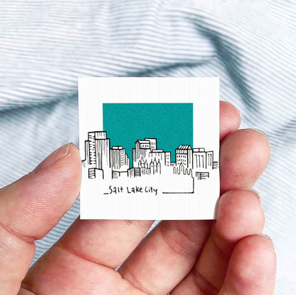 A hand holding a small square-shaped object with a drawing of a city skyline and the text "Salt Lake City" written on it.