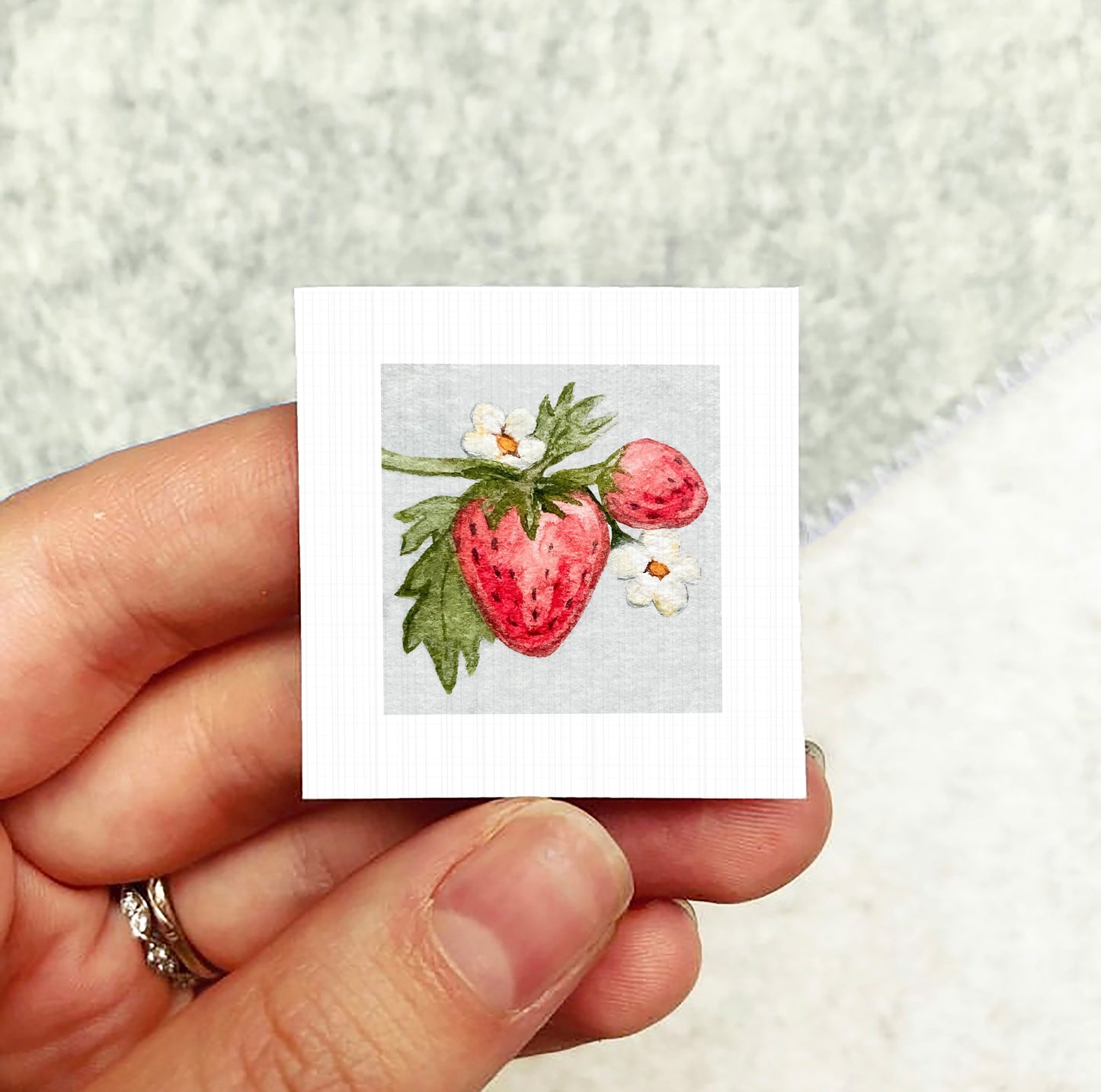 A hand holding a small square card with a painting of a strawberry and white flowers on it.