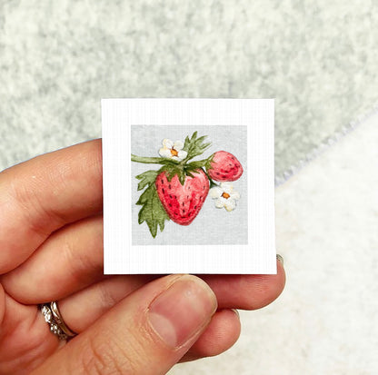 A hand holding a small square card with a painting of a strawberry and white flowers on it.
