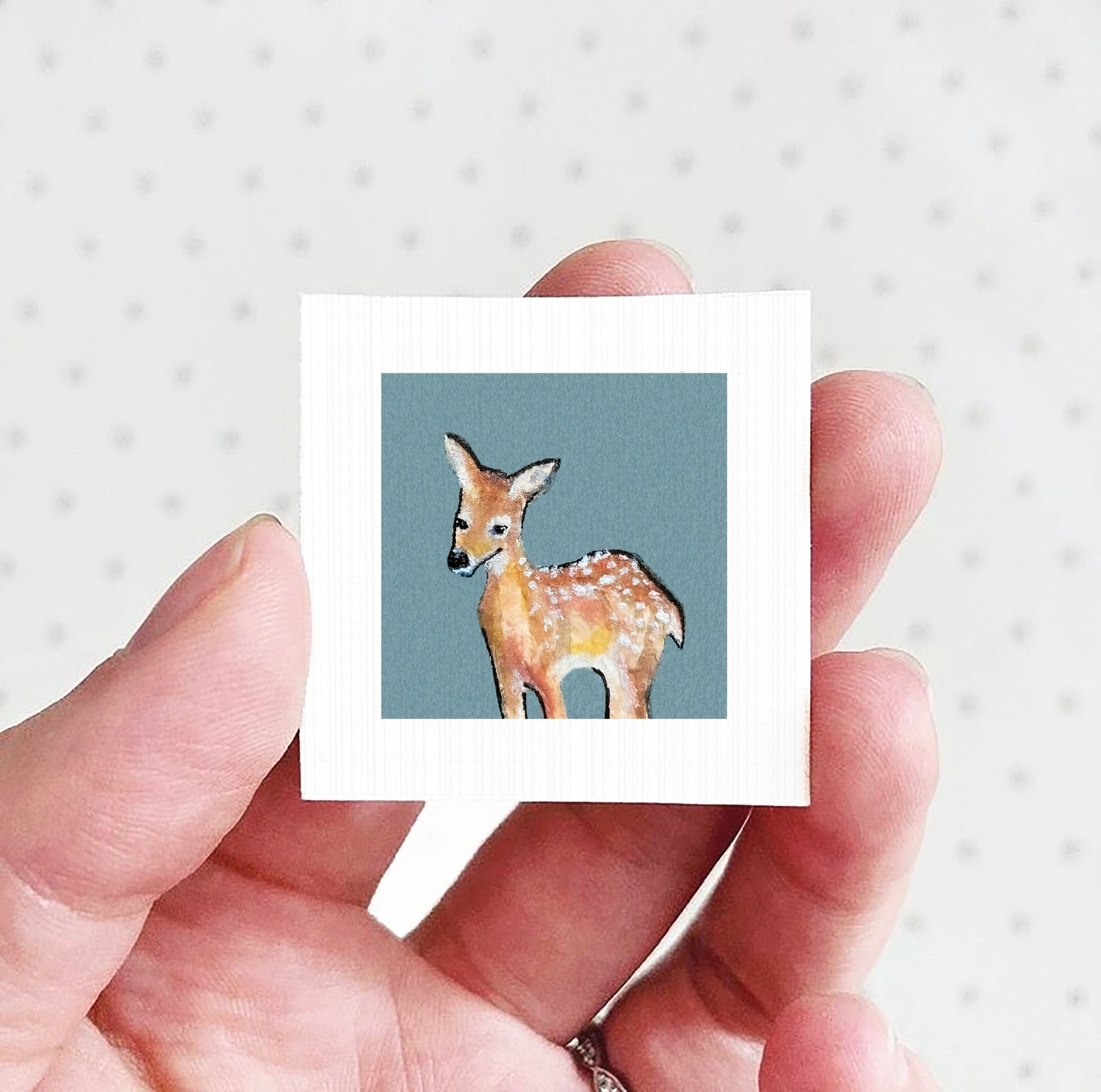 A hand holding a small square picture of a deer against a light blue background.