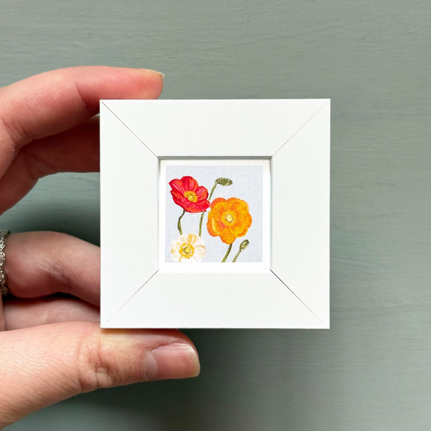 A hand is holding a small white frame containing a painting of two flowers, one red and one yellow.