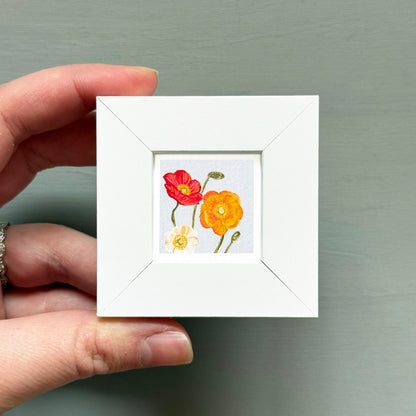 A hand is holding a small white frame containing a painting of two flowers, one red and one yellow.