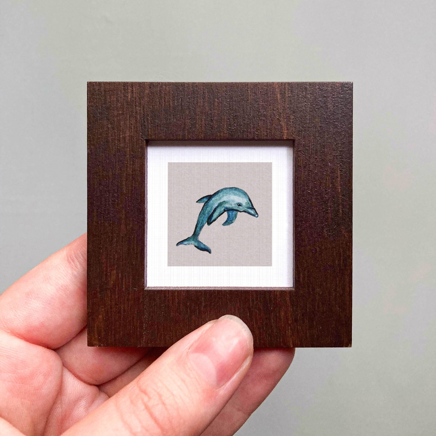 A hand is holding a small wooden frame containing a watercolor painting of a dolphin.