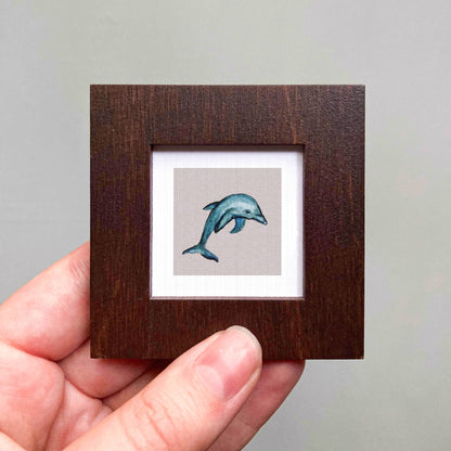 A hand is holding a small wooden frame containing a watercolor painting of a dolphin.