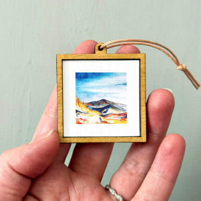 A hand holding a small wooden frame containing a watercolor painting of a mountain landscape.