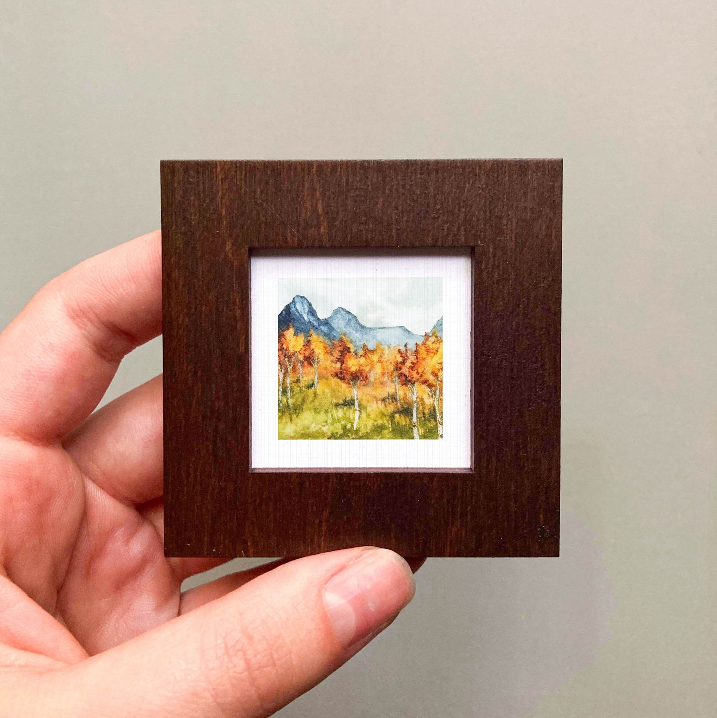 A hand is holding a small, square-shaped wooden frame containing a watercolor painting of a mountain landscape with autumn trees.