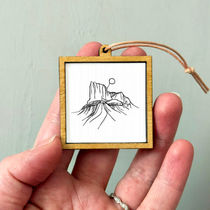 A hand holding a small wooden frame containing a black and white line drawing of a mountain landscape.