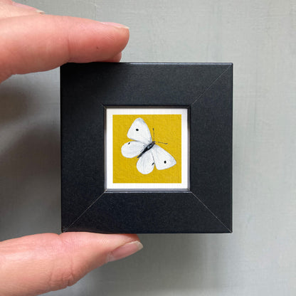 A person's hand is holding a small black frame containing a framed image of a white butterfly on a yellow background.