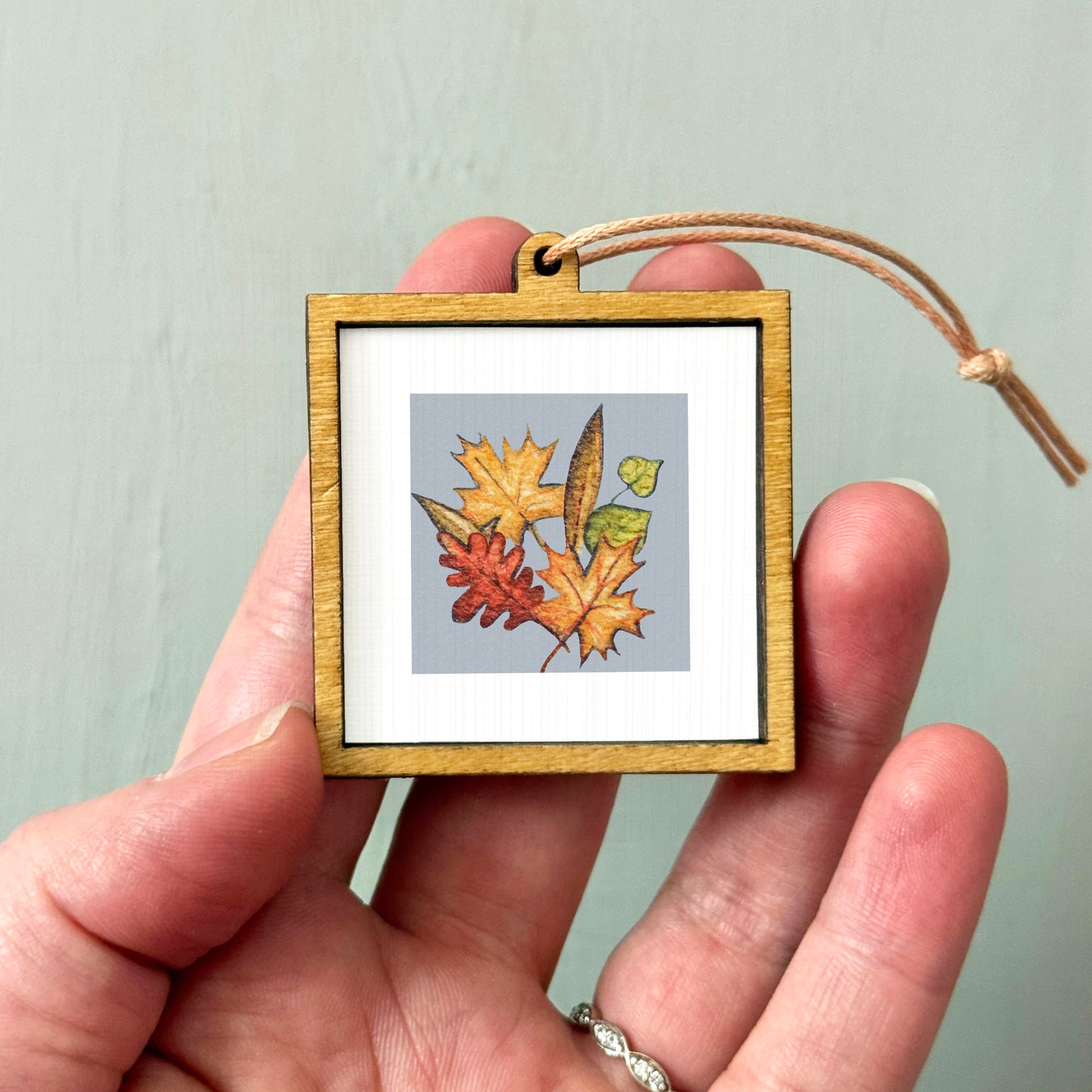 A hand holding a small wooden frame containing a framed image of autumn leaves.
