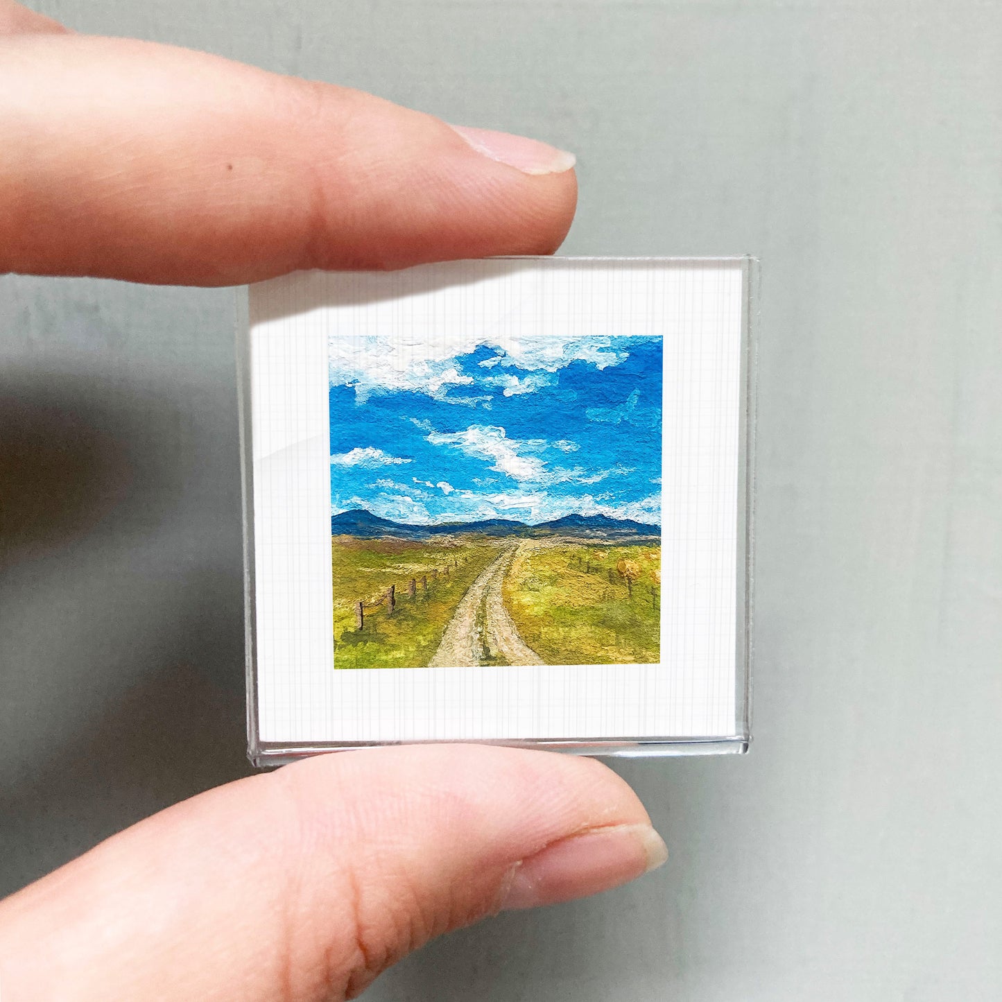A person's hand holding a small square picture of a rural landscape with a dirt road leading through a grassy field and mountains in the background.