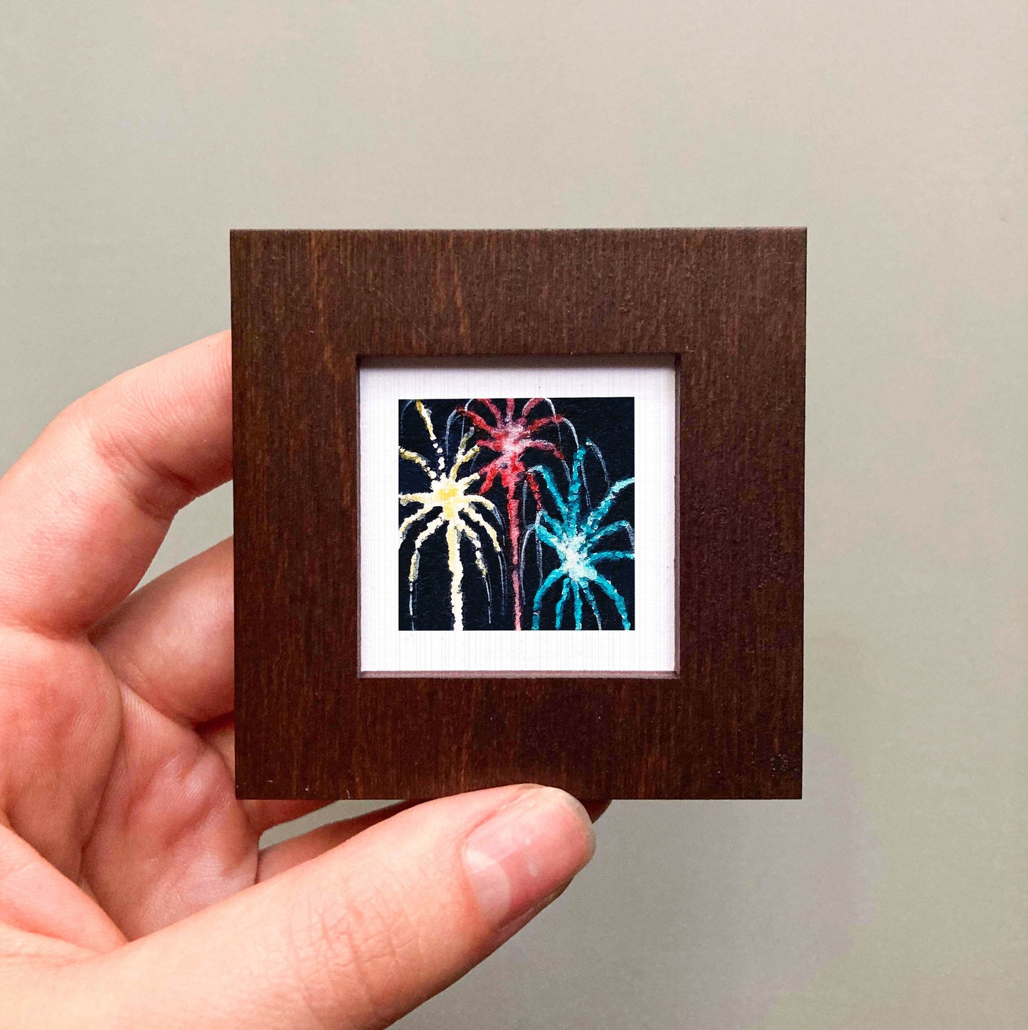 A hand is holding a small wooden frame containing a colorful painting of fireworks.