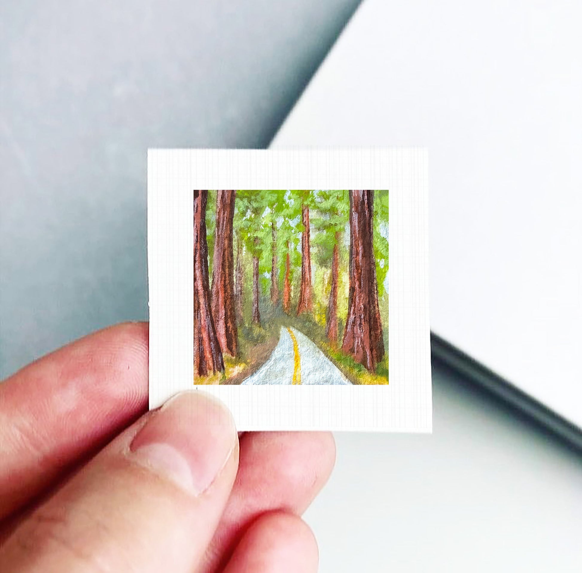 A hand is holding a small, framed painting of a forest road with tall trees on either side.