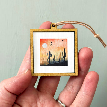 A hand holding a small wooden frame containing a painting of cacti silhouetted against a sunset sky.