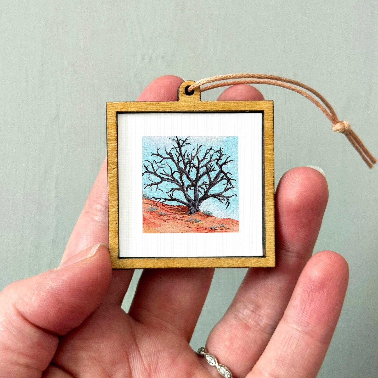 A hand holding a small wooden frame containing a painting of a bare tree against a blue sky.