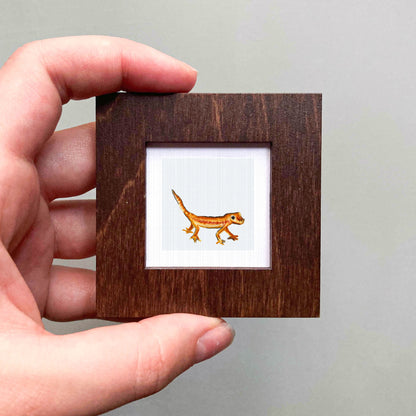 A hand is holding a small wooden frame containing a picture of a lizard.