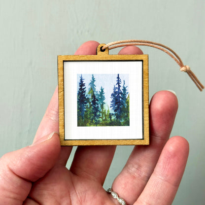 A hand holding a small wooden frame containing a painting of a forest scene.