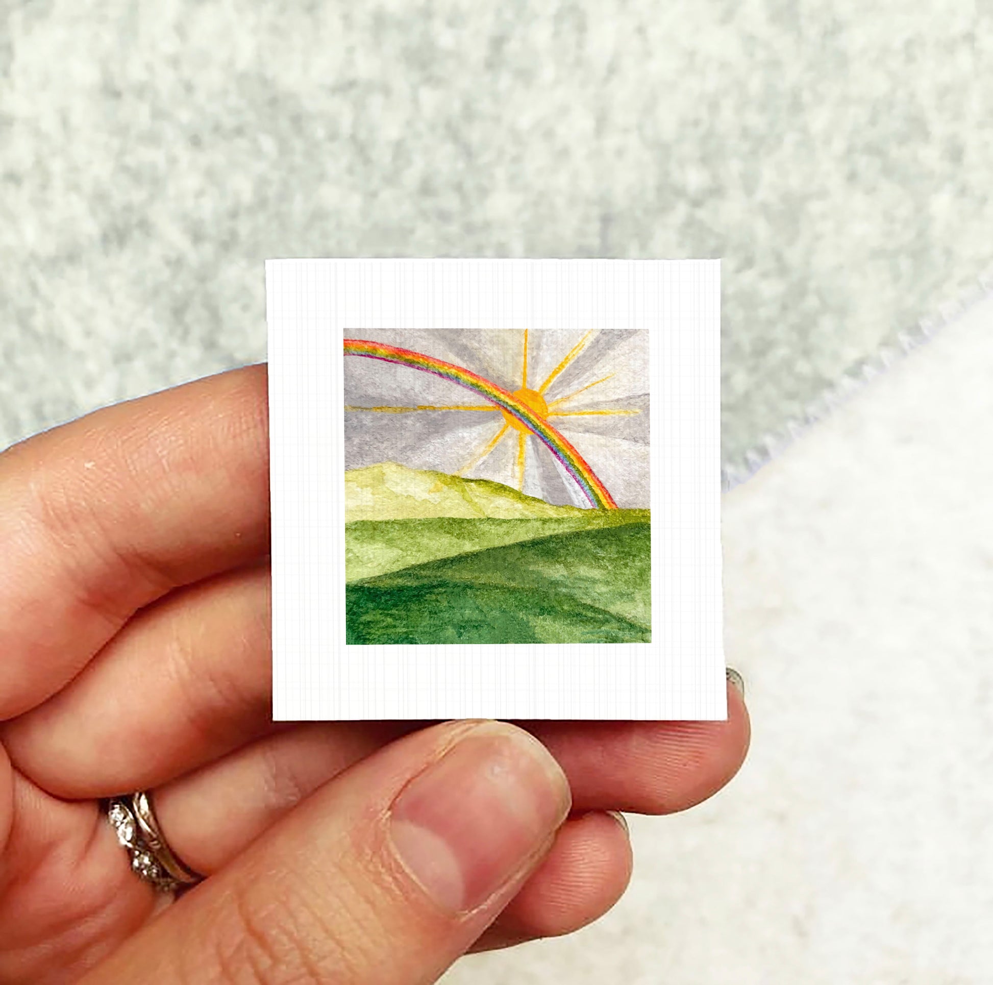 A hand holding a small square artwork depicting a vibrant rainbow over a lush green landscape.