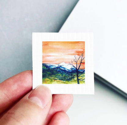 A hand is holding a small square picture depicting a landscape with mountains and a tree.