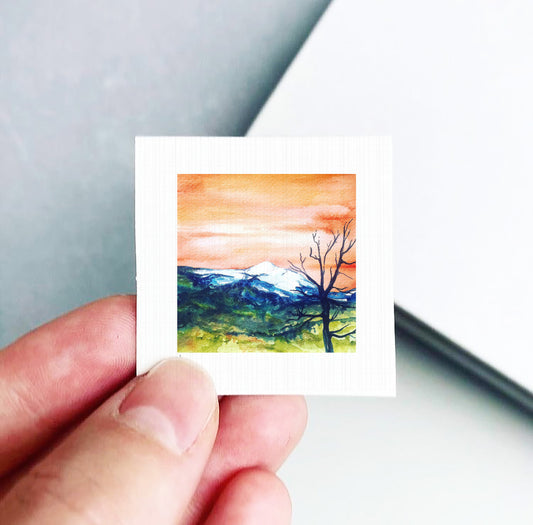 A hand is holding a small square picture depicting a landscape with mountains and a tree.