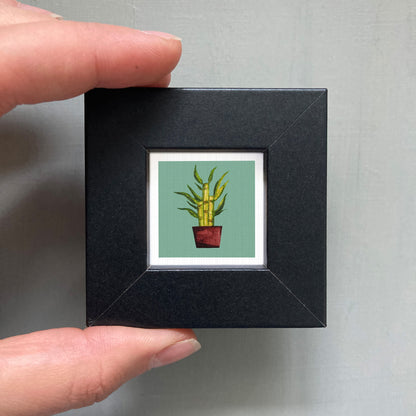 A person's hand holding a small black frame containing a framed illustration of a potted plant.