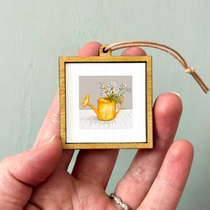 A hand holding a small wooden frame containing a painting of a yellow watering can with flowers.