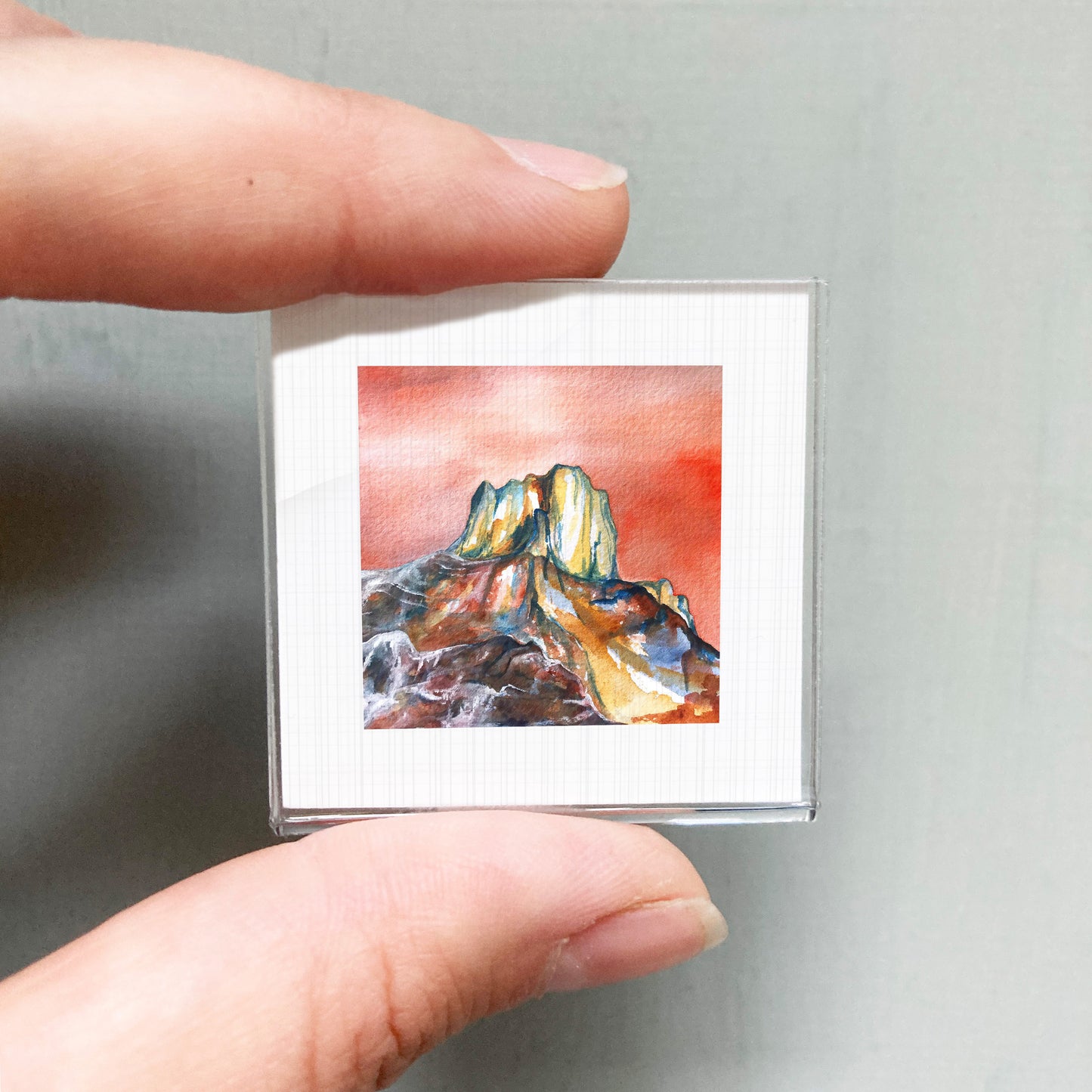 A person's hand holding a small square picture of a mountain landscape with a red and orange background.