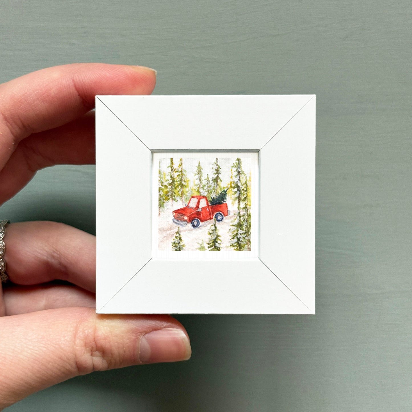 A hand is holding a small white picture frame containing a painting of a red truck driving through a snowy forest.