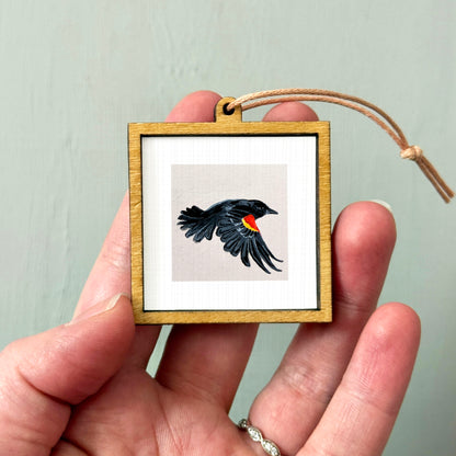A person's hand holding a small wooden frame containing a framed image of a black bird in flight.