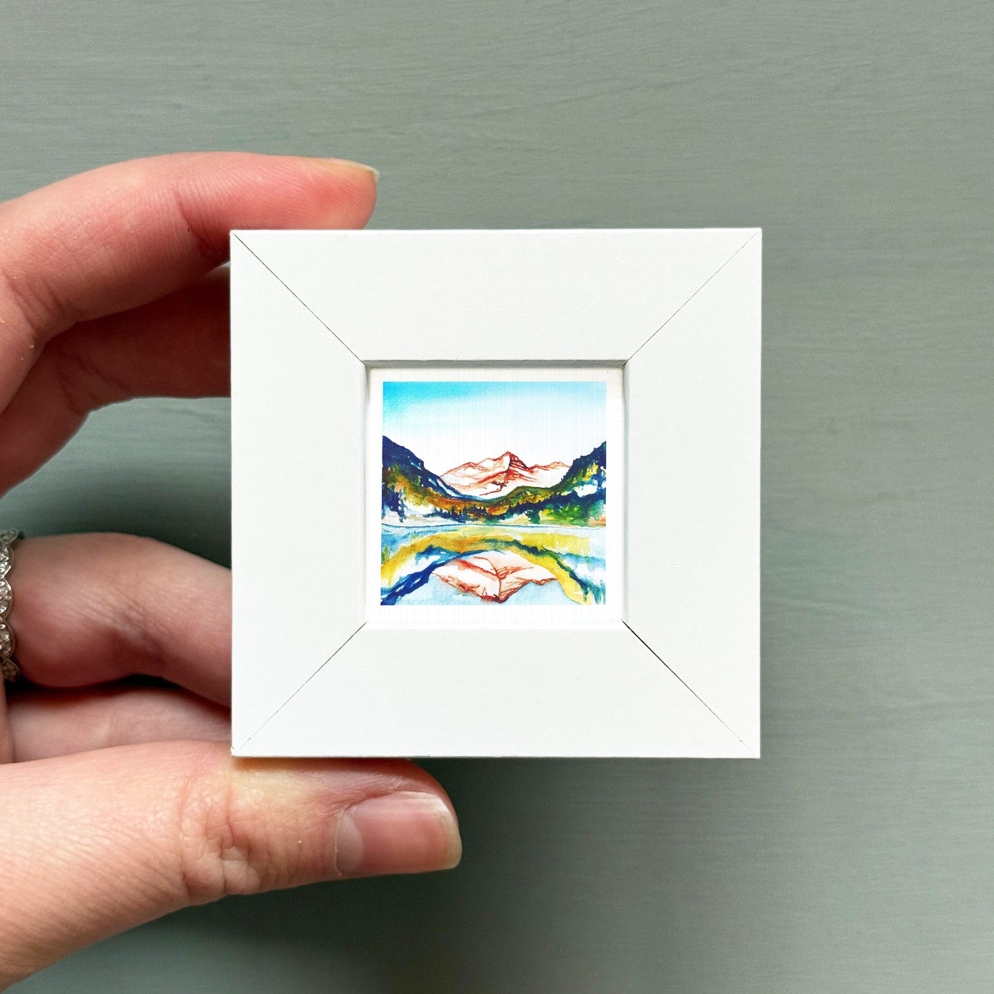 A hand is holding a small white square frame containing a watercolor painting of a mountain landscape.