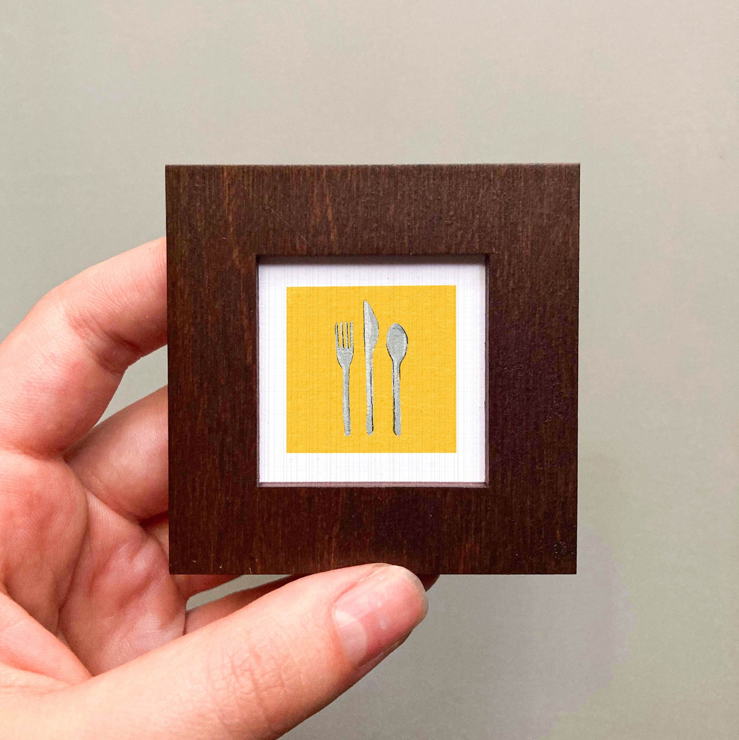 A hand is holding a small wooden frame containing a framed image of three utensils - a fork, a knife, and a spoon - against a yellow background.