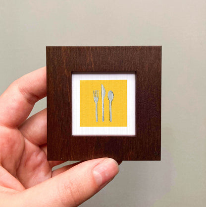 A hand is holding a small wooden frame containing a framed image of three utensils - a fork, a knife, and a spoon - against a yellow background.