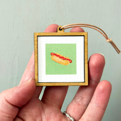 A person's hand holding a small wooden frame containing a picture of a hot dog.