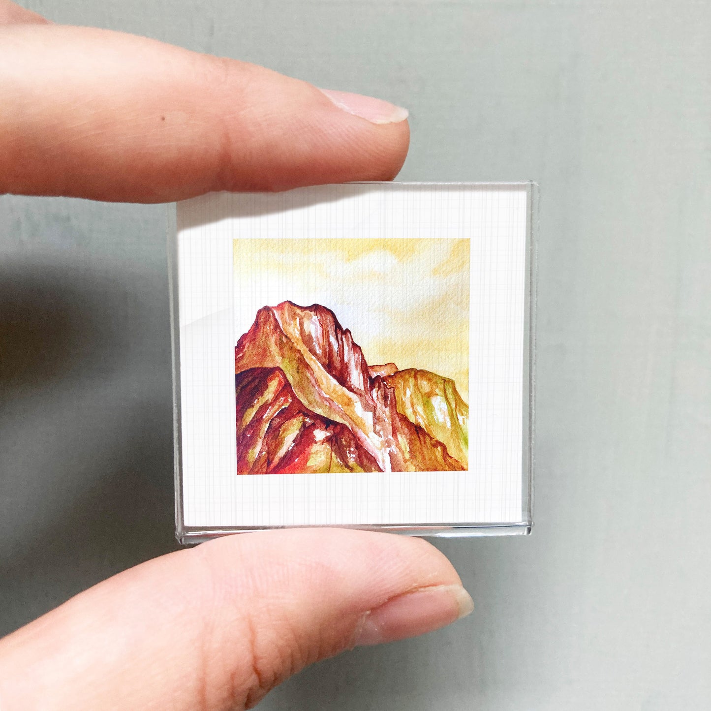 A person's hand holding a small square picture of a mountain landscape.