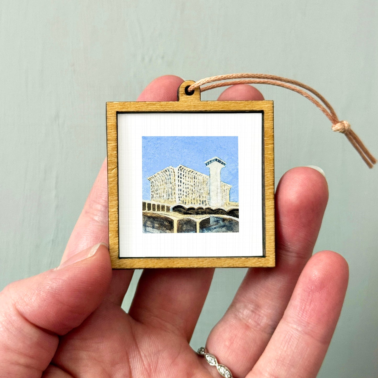 A hand holding a small wooden frame containing a miniature painting of a building.