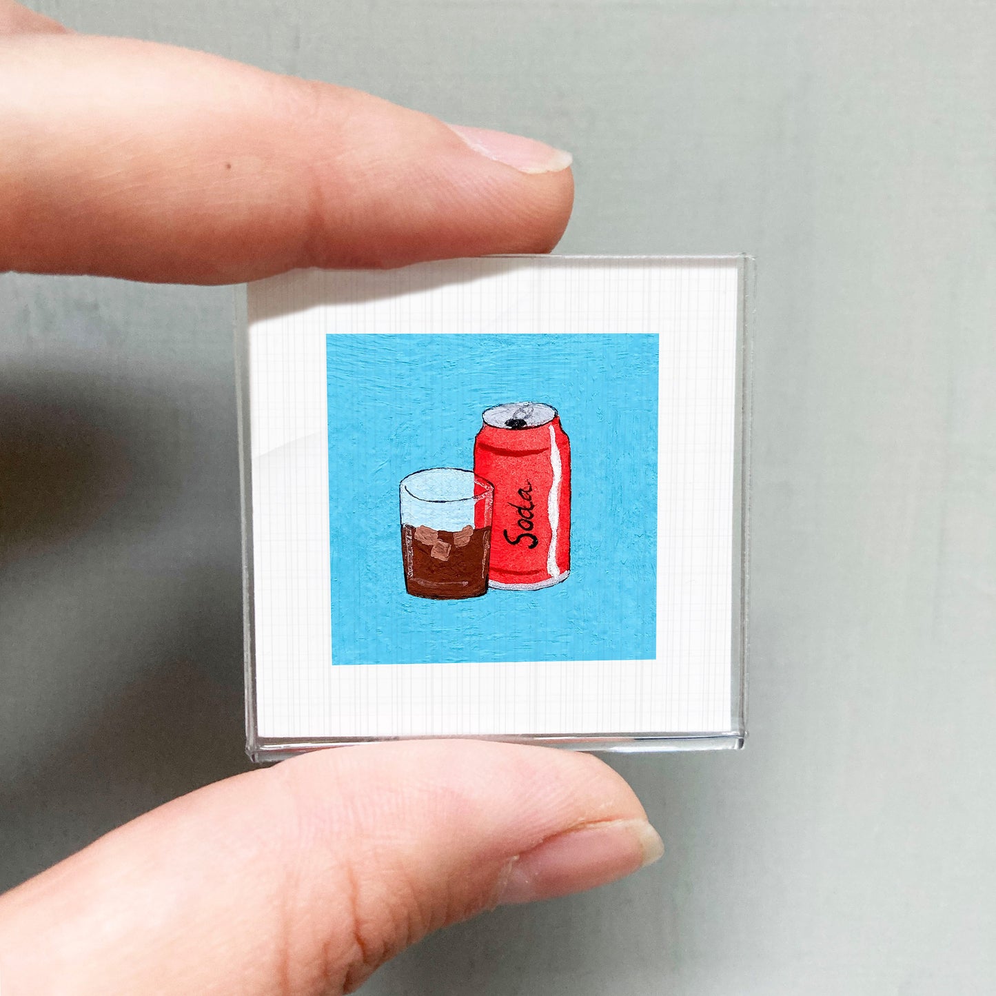 A person's hand holding a small square with an illustration of a glass of soda and a can of soda on it.