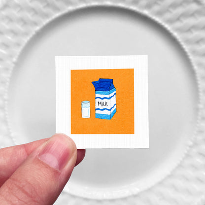 A hand is holding a small square with an illustration of a carton of milk and a salt shaker on it.