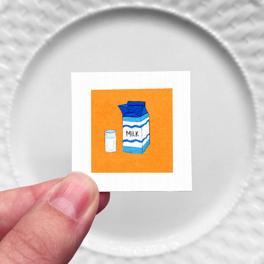 A hand is holding a small square with an illustration of a carton of milk and a salt shaker on it.